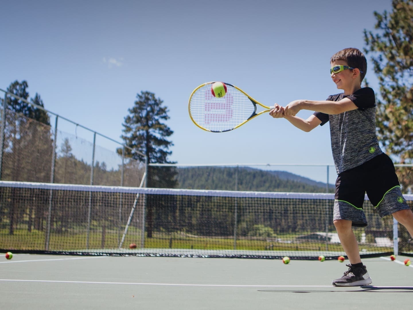 Little boy hitting a tennis ball with a racquet on a court in the mountains