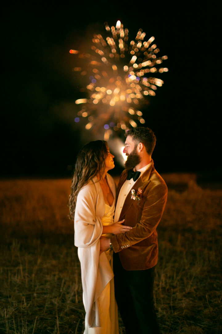 Bride and groom outside at night with fireworks behind them.