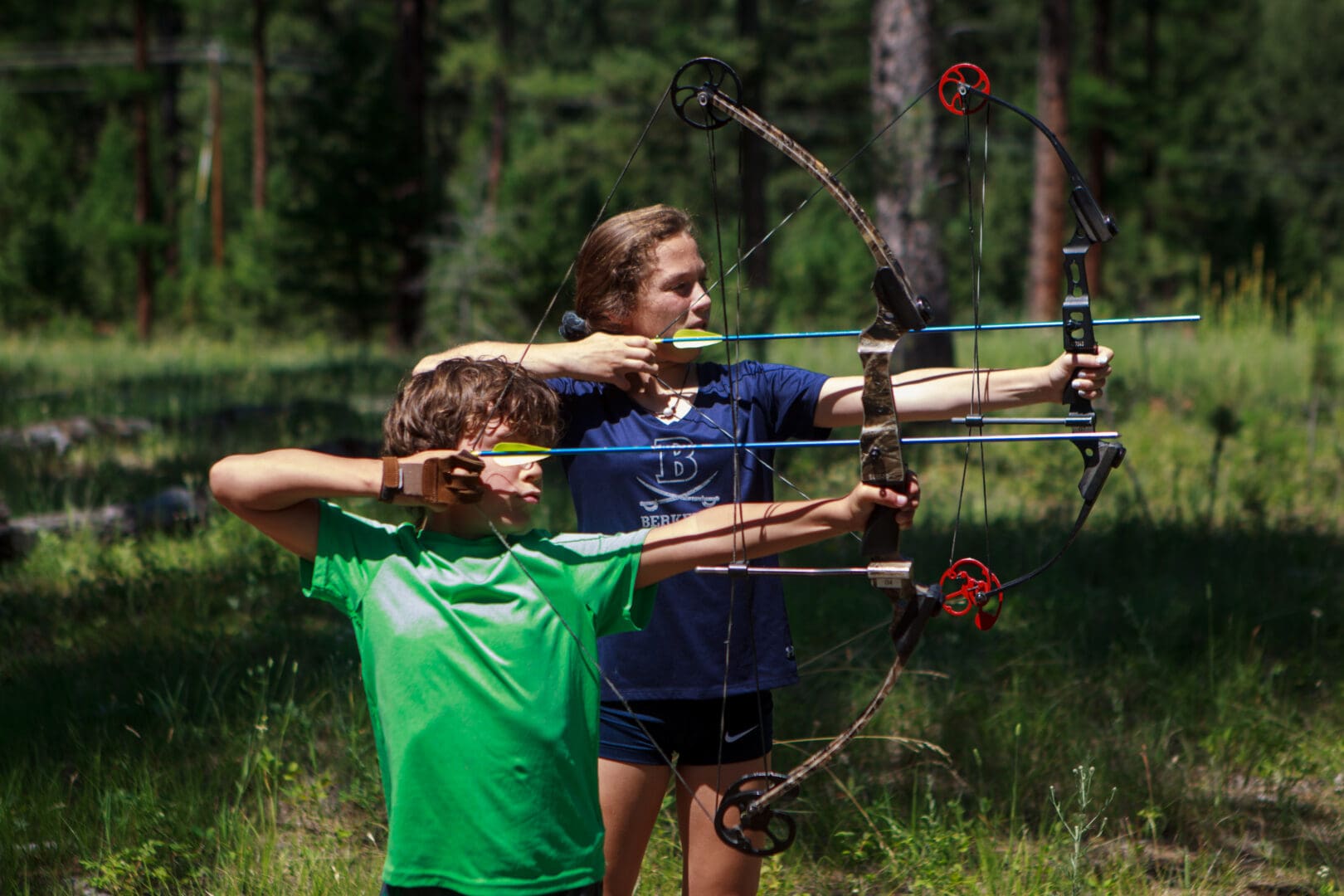 Girl and boy shooting a bow and arrow in a forest.