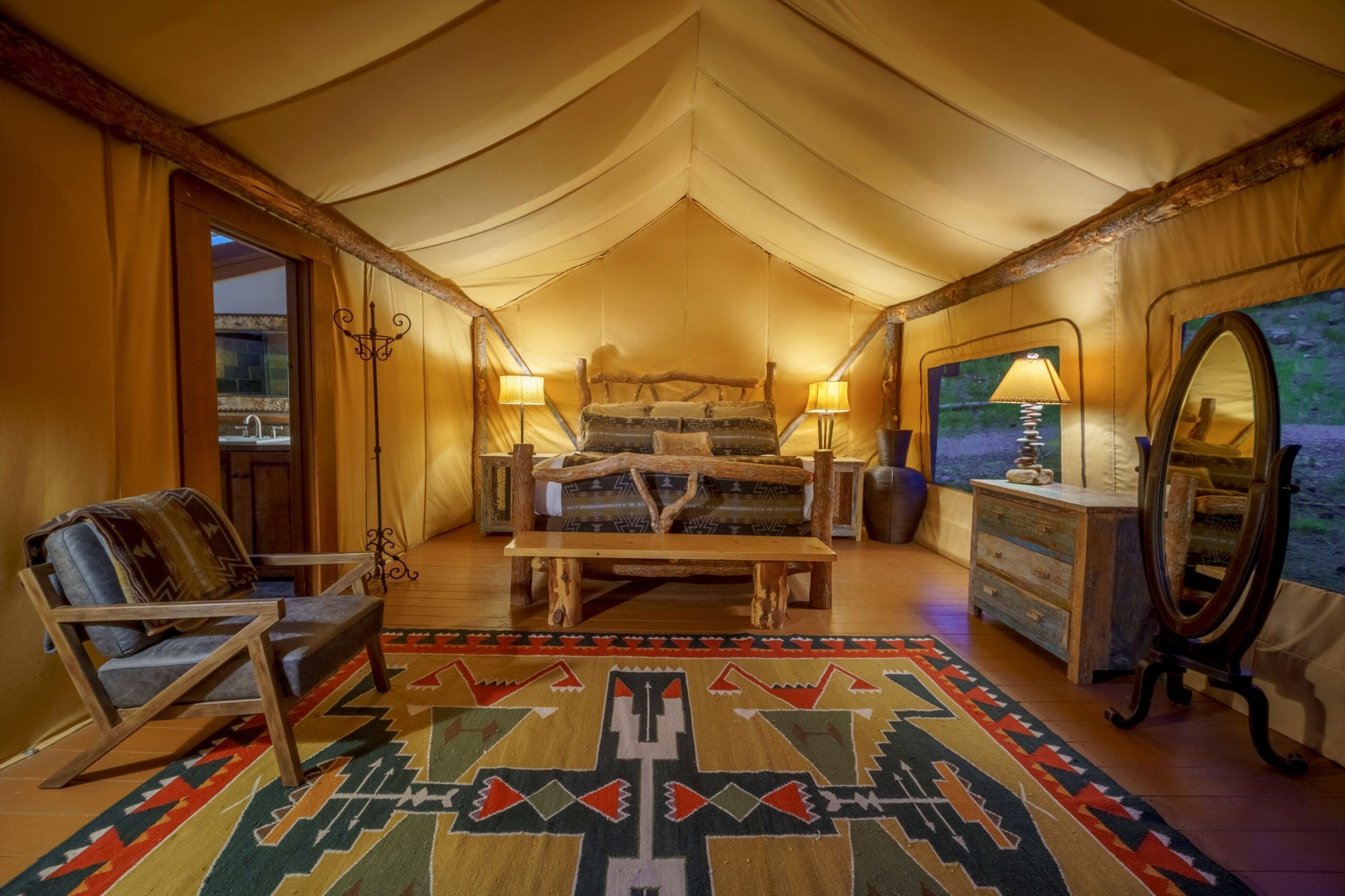 Inside of a yellow Paws Up glamping tent, featuring a luxury, king-size bed, leather and wooden armchair, warm, canvas lamps, and an aerial rug
