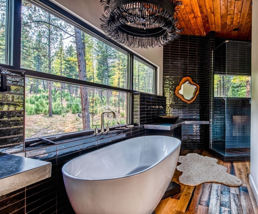 Bathroom in the Tree Haus at Green O, featuring dark tiled wall, a bathtub under a modern chandelier, and a bright forest view from the window
