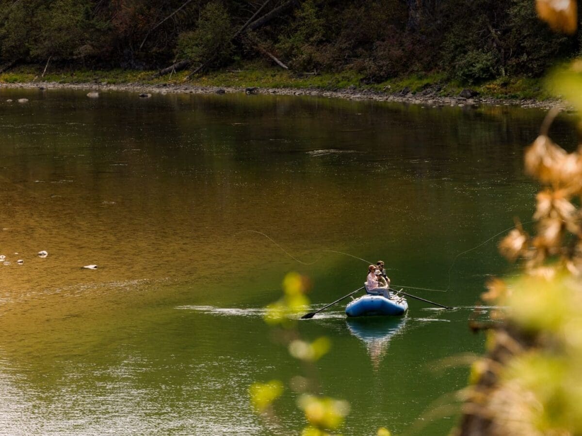 Wide view of a group of people paddling a blue raft through the Blackfoot River to fly fish
