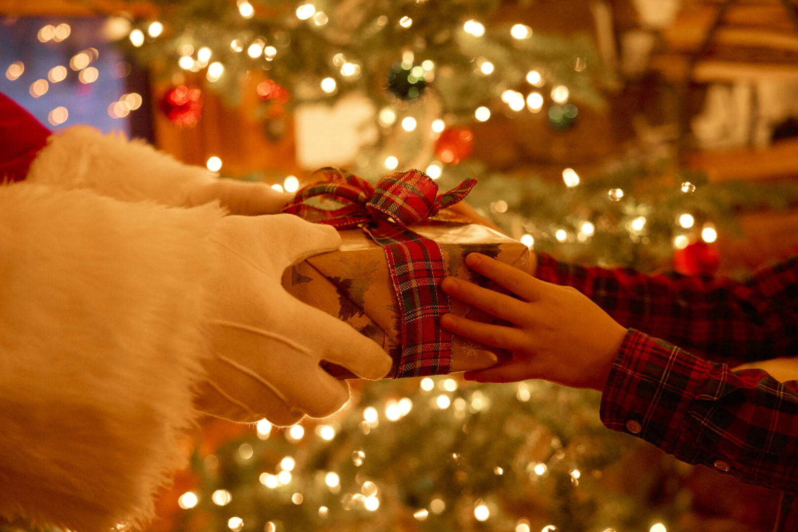 Santa Claus handing a child a present wrapped in a bow with a Christmas tree behind them.