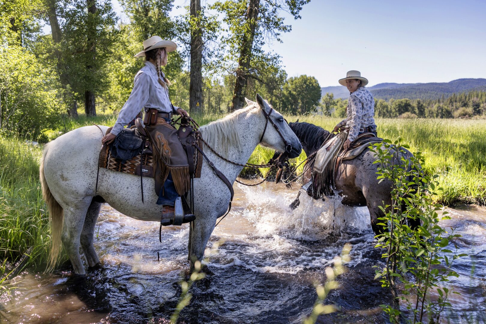 Two wranglers on their horses standing in a creek with trees behind them.