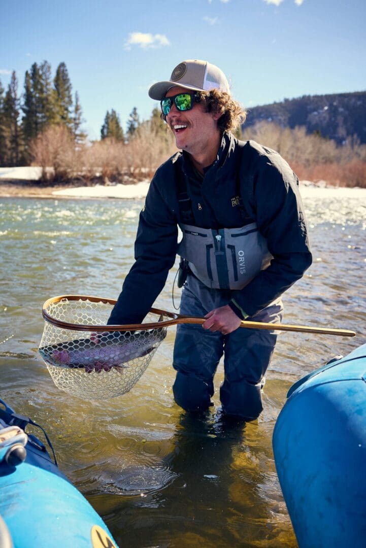 Fisher wearing waders in a river during the winter, holding a fish in a net.