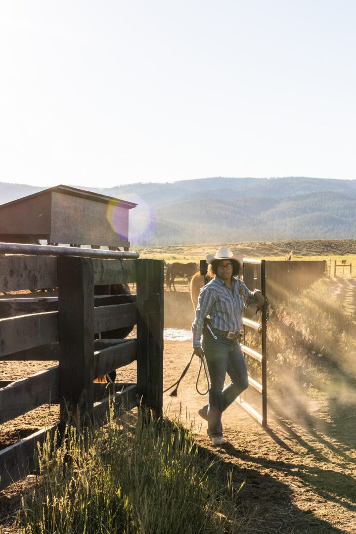 Woman wrangler opening up a wooden gate for horses.