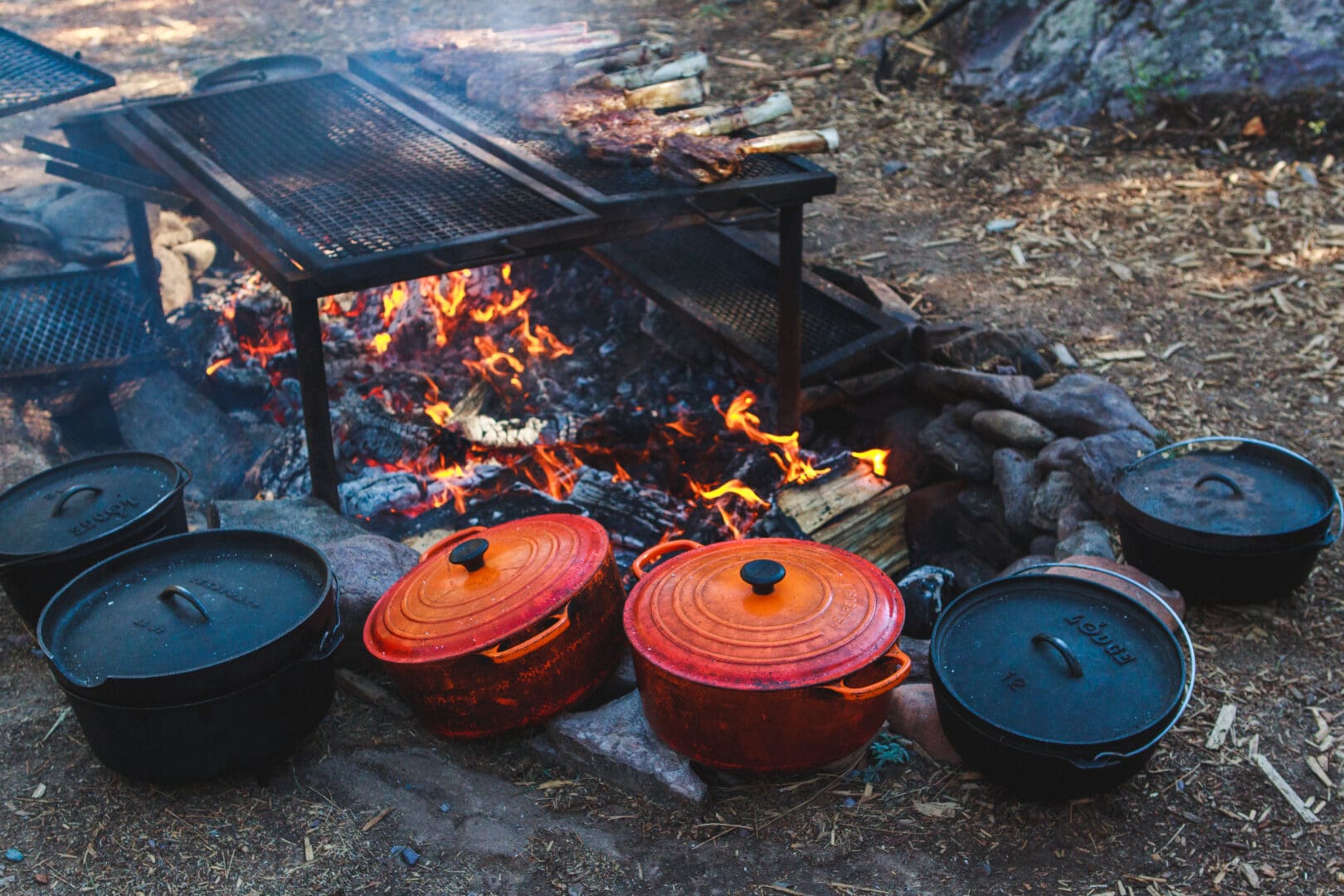 Several red and white dutch ovens cooking food around a fire.