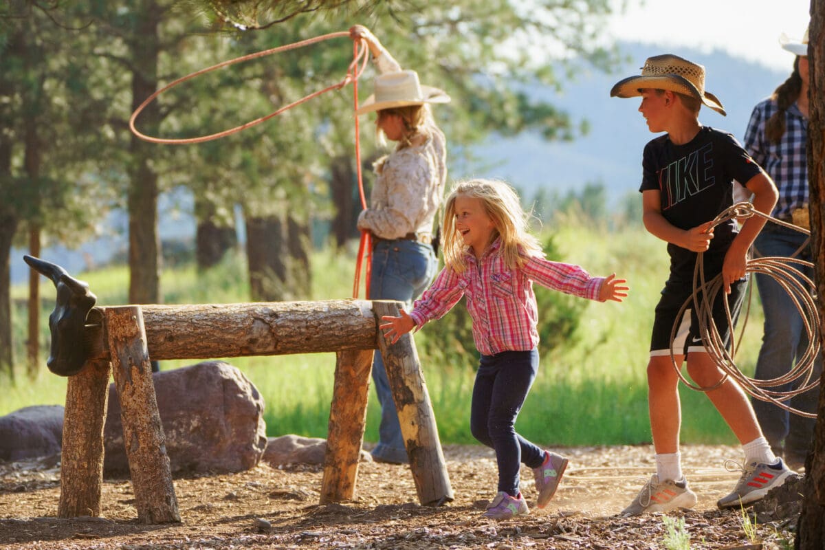 Two children running around a western land while a lady tries to rope a fake cattle.