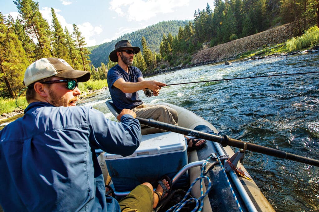 Two men in a raft on a river, one rowing and one fishing.