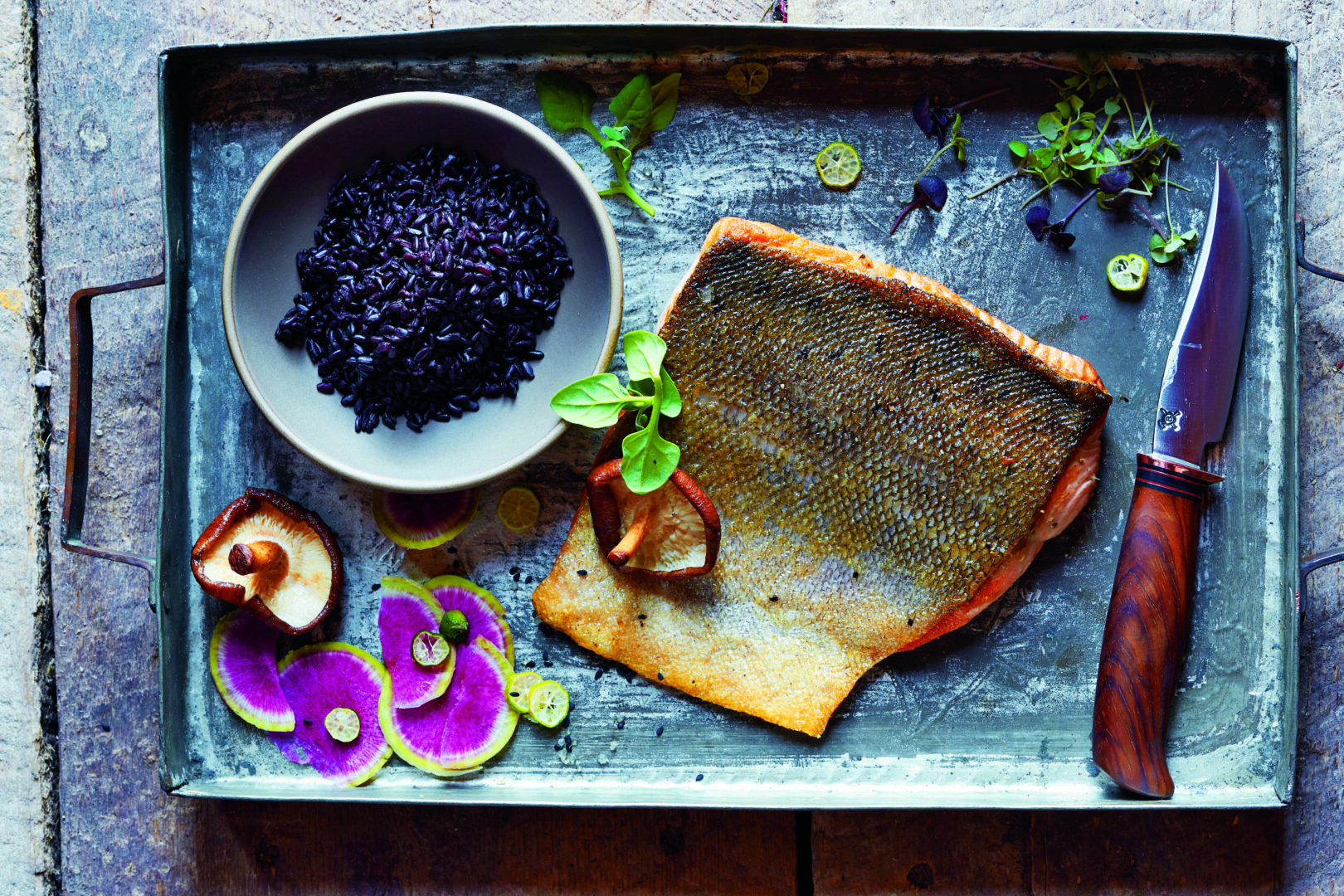 Cavier in a bowl, cooked fish, and radishes next to a knife, all laid out on a rectangular dinner platter.