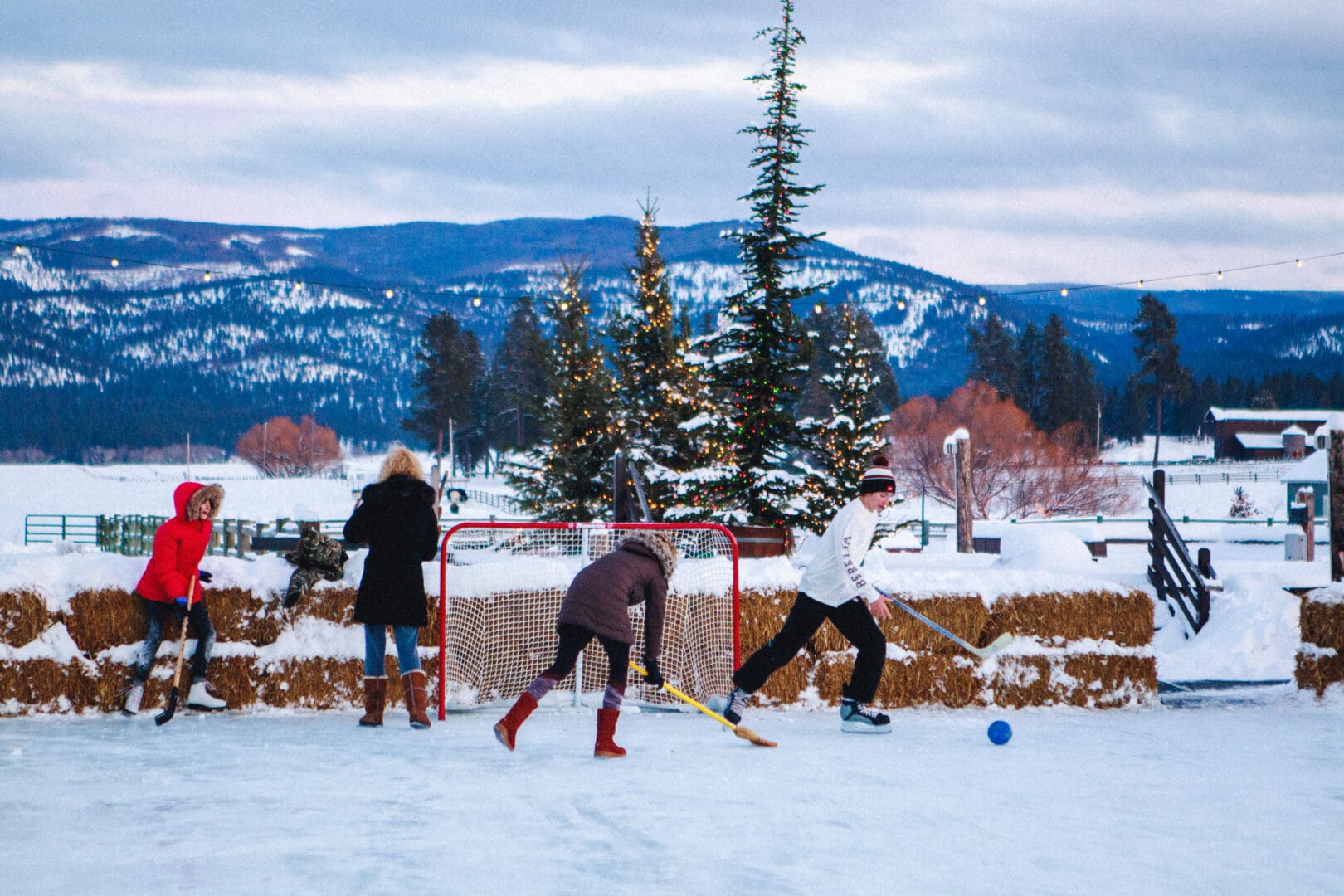 Large group or family playing hockey on an outdoor ice rink with snow and mountains in the background.