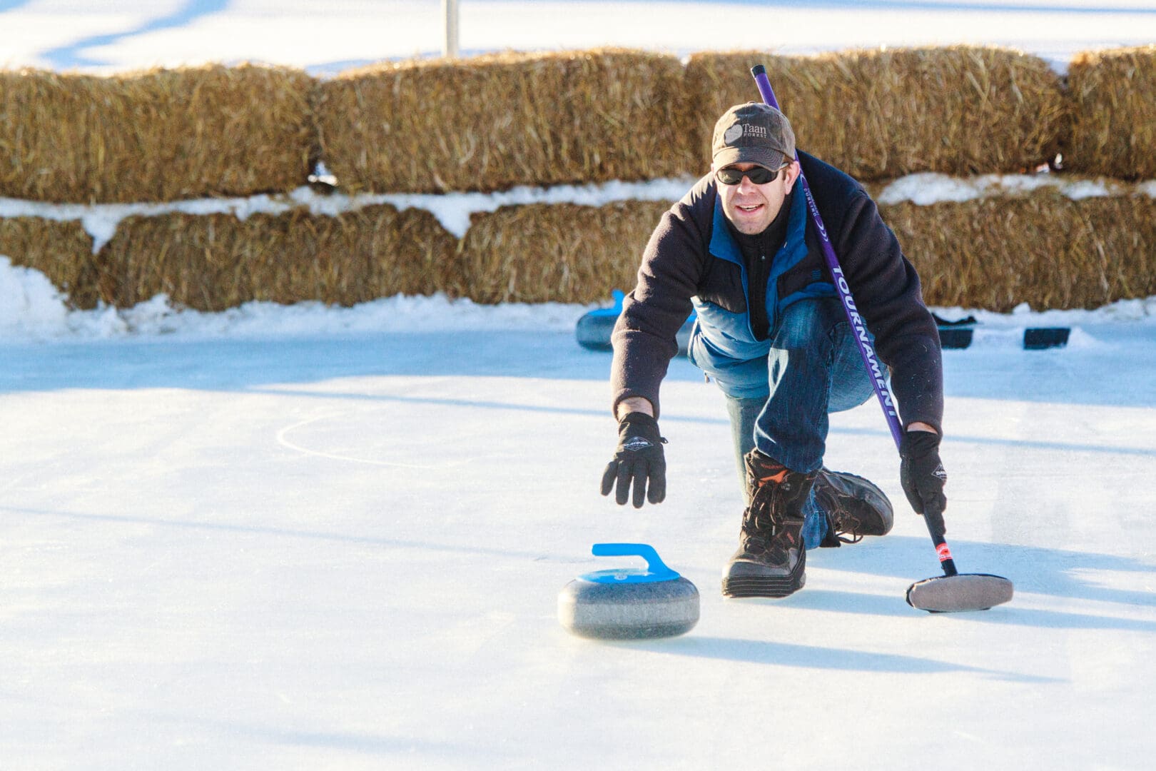 Man on ice playing curling with stack of hay behind him.