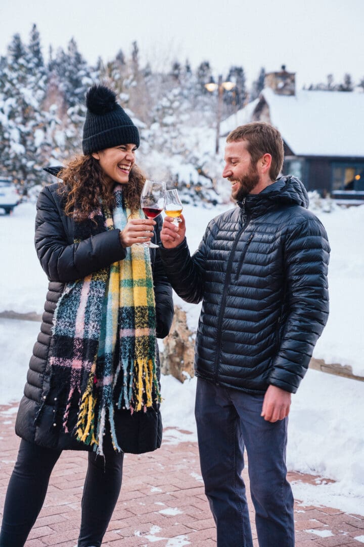 Couple in winter jackets outside standing in the snow, clinking glasses together.