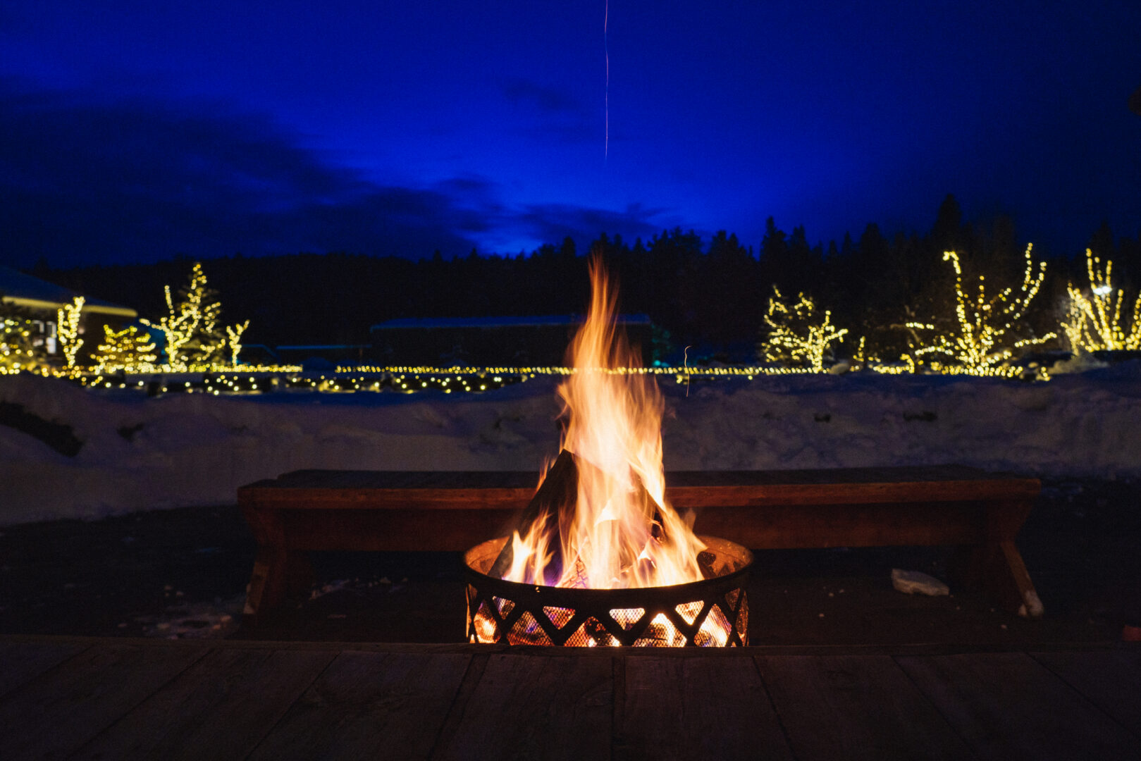 A fire at night in the winter with lights covering trees in the distance.