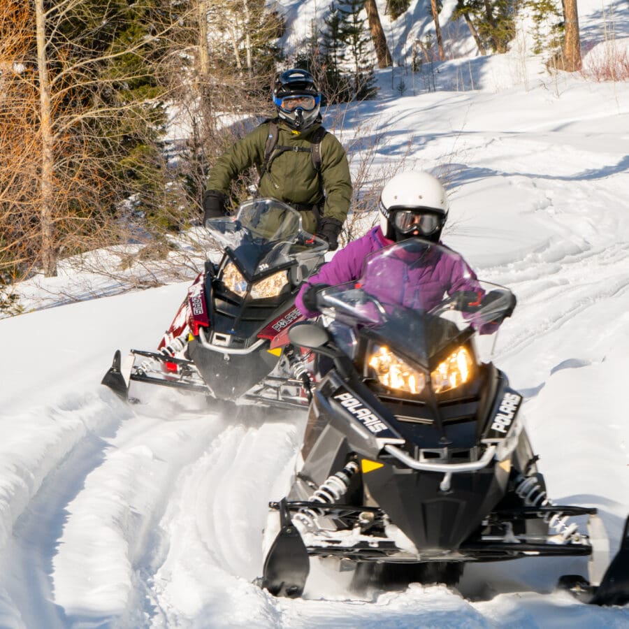 Two people snowmobiling in gear outside in the snow.