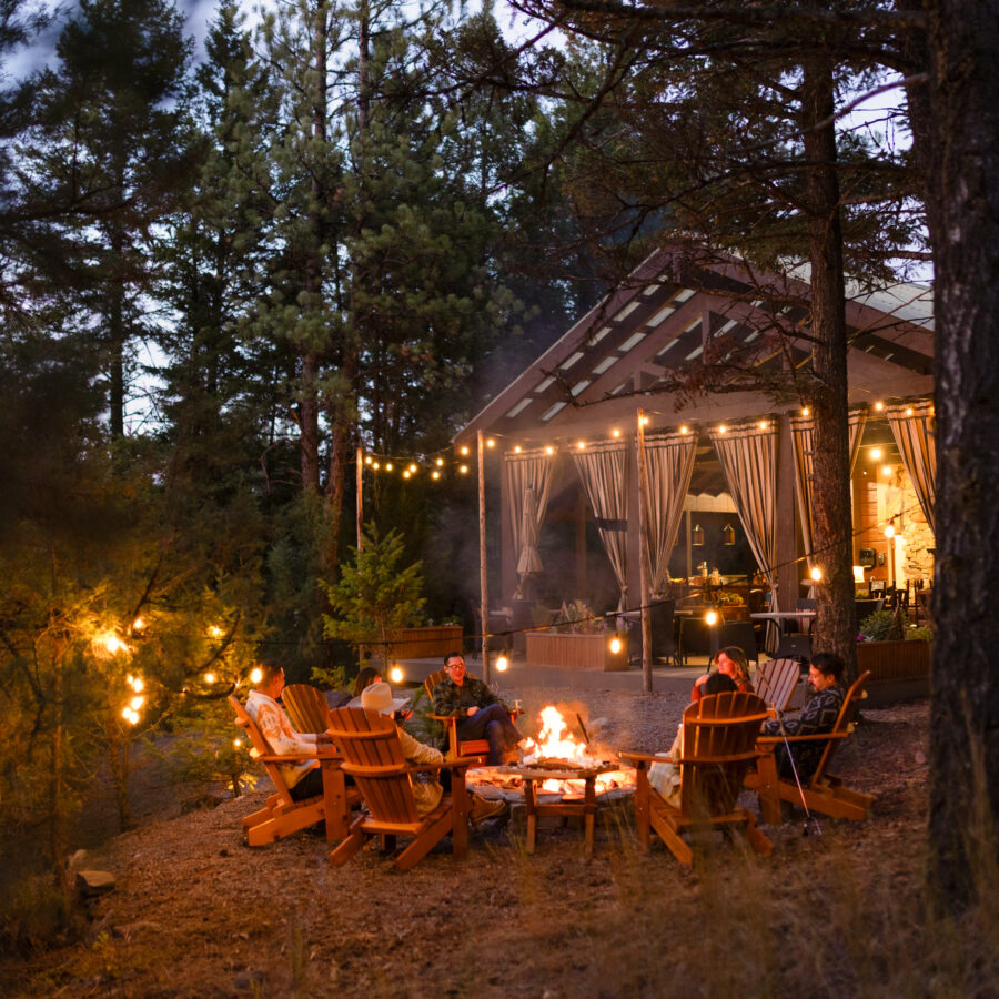 Group of individuals sitting around a fire at sunset with lights and a light up pavilion behind them.