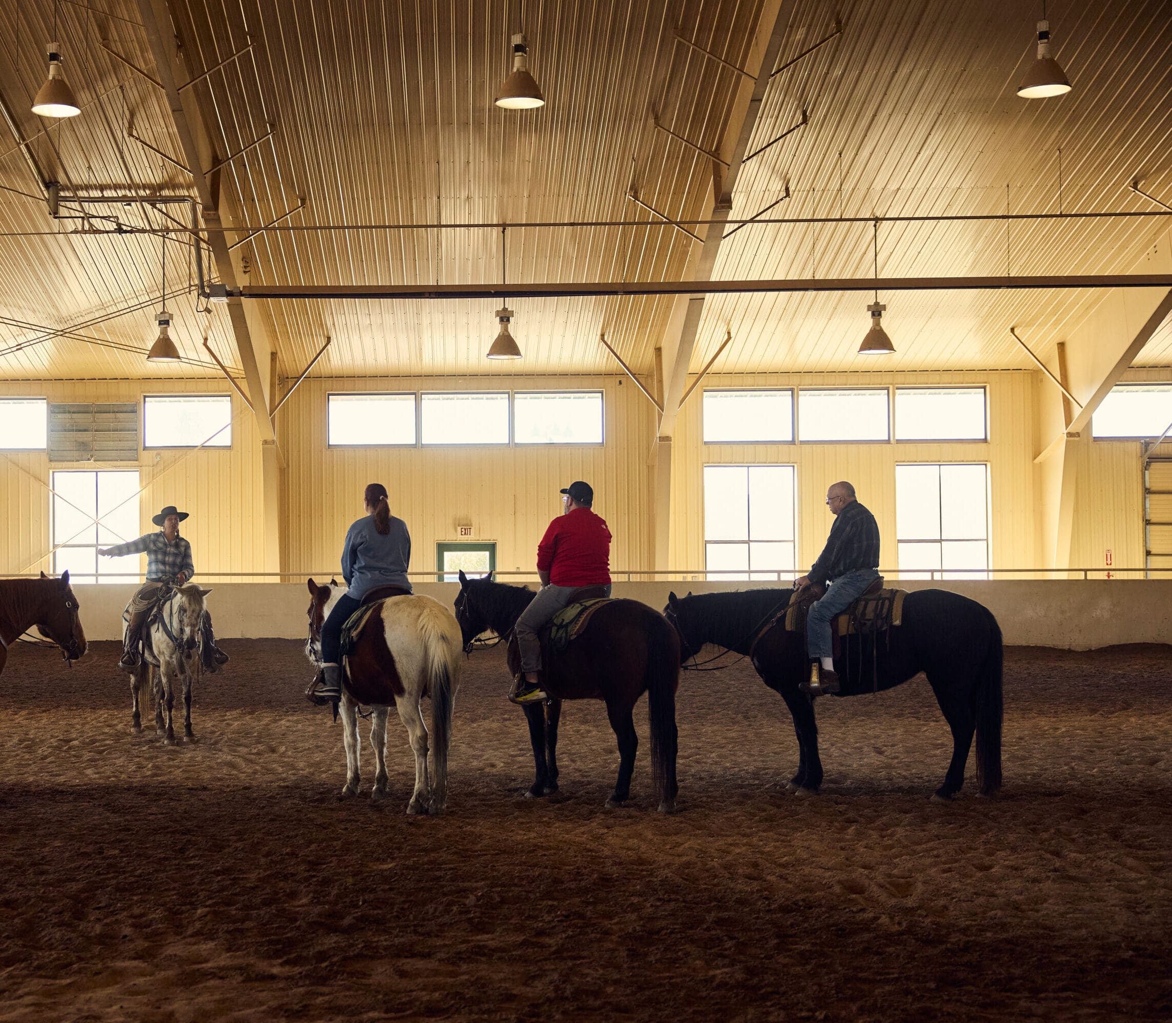 Group of individuals sitting on horses in an arena, while an instructor talks to them.