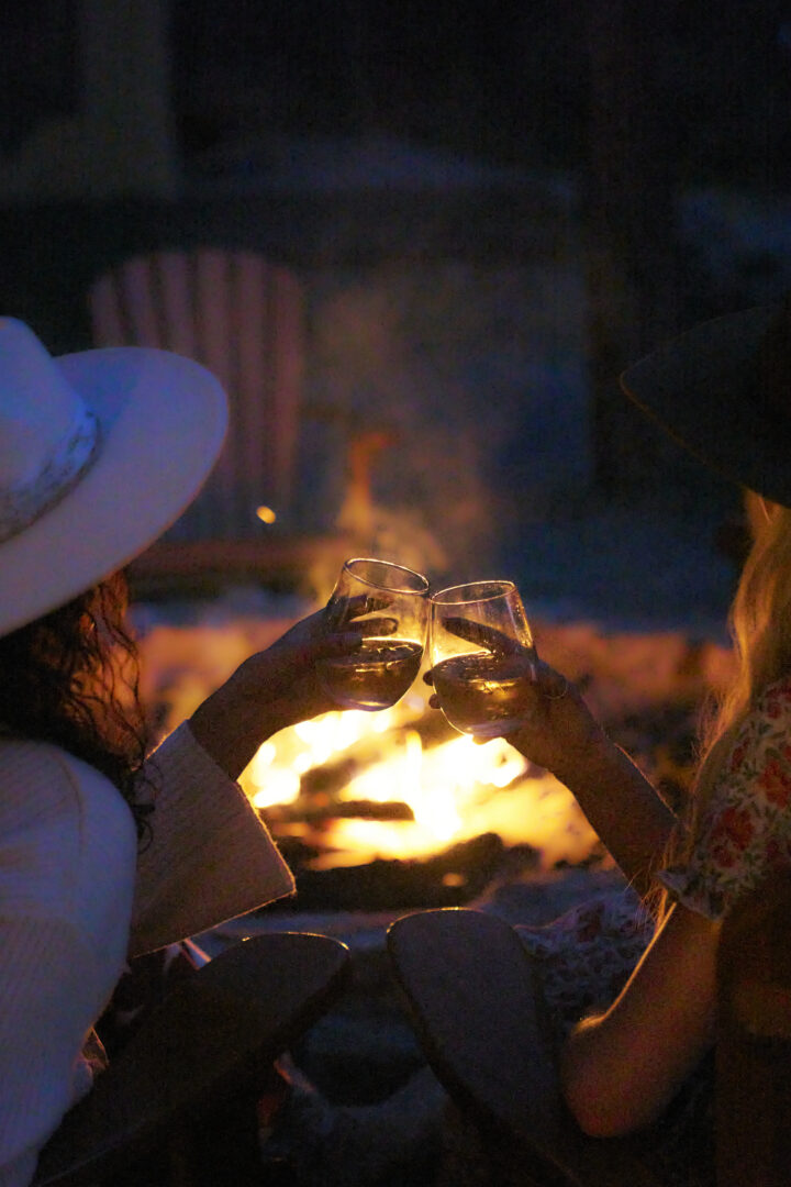 Two women sitting at a fire clinking glasses of wine.