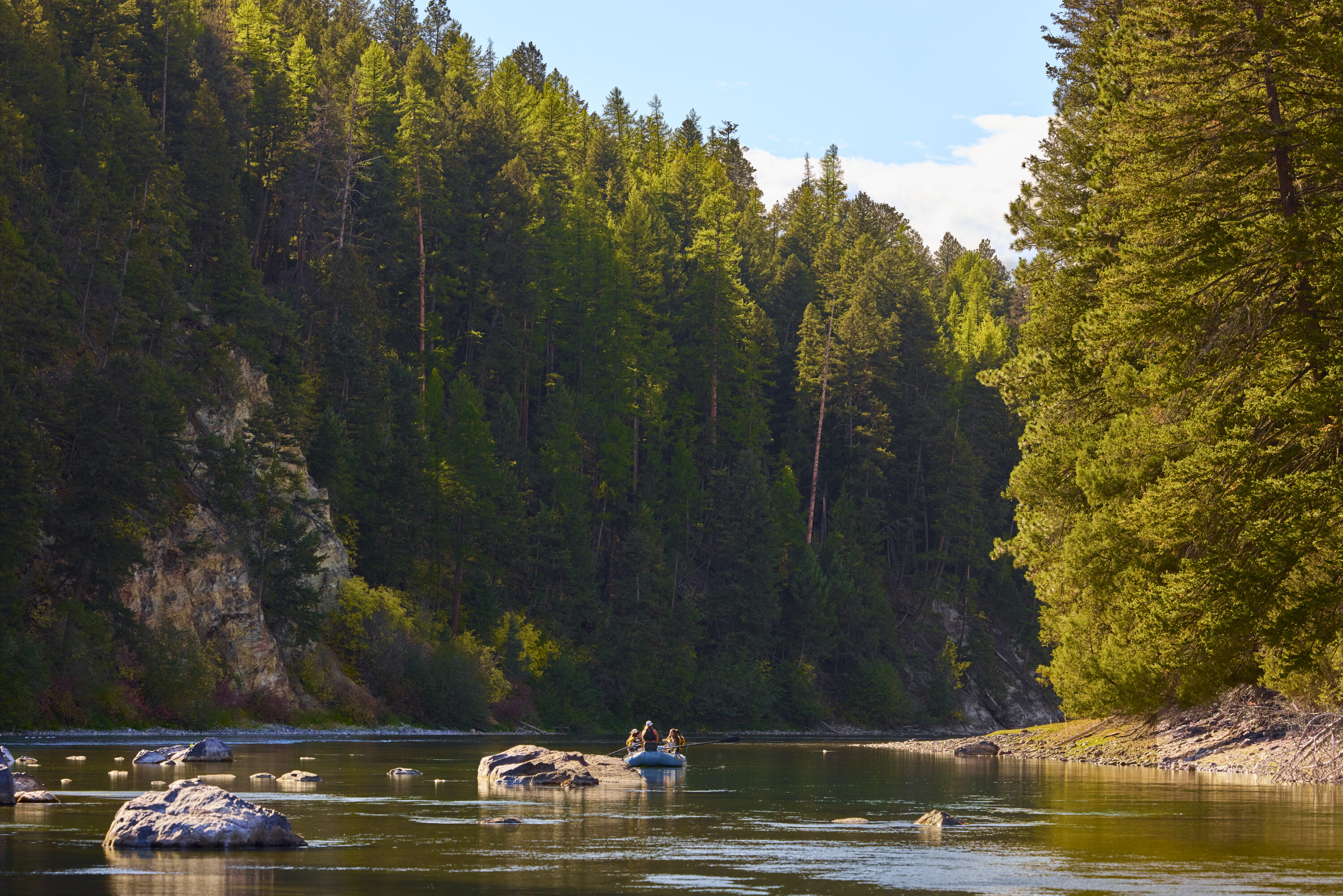 Shot of a river with trees and a small blue boat with people fly fishing.