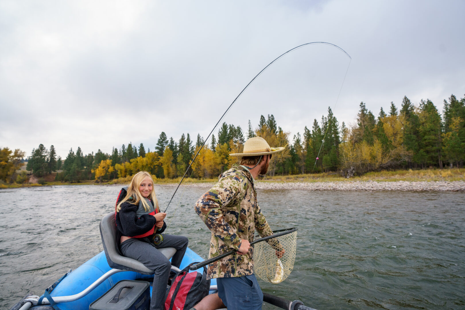 Man and young girl fishing in a blue boat with trees behind them in the distance.