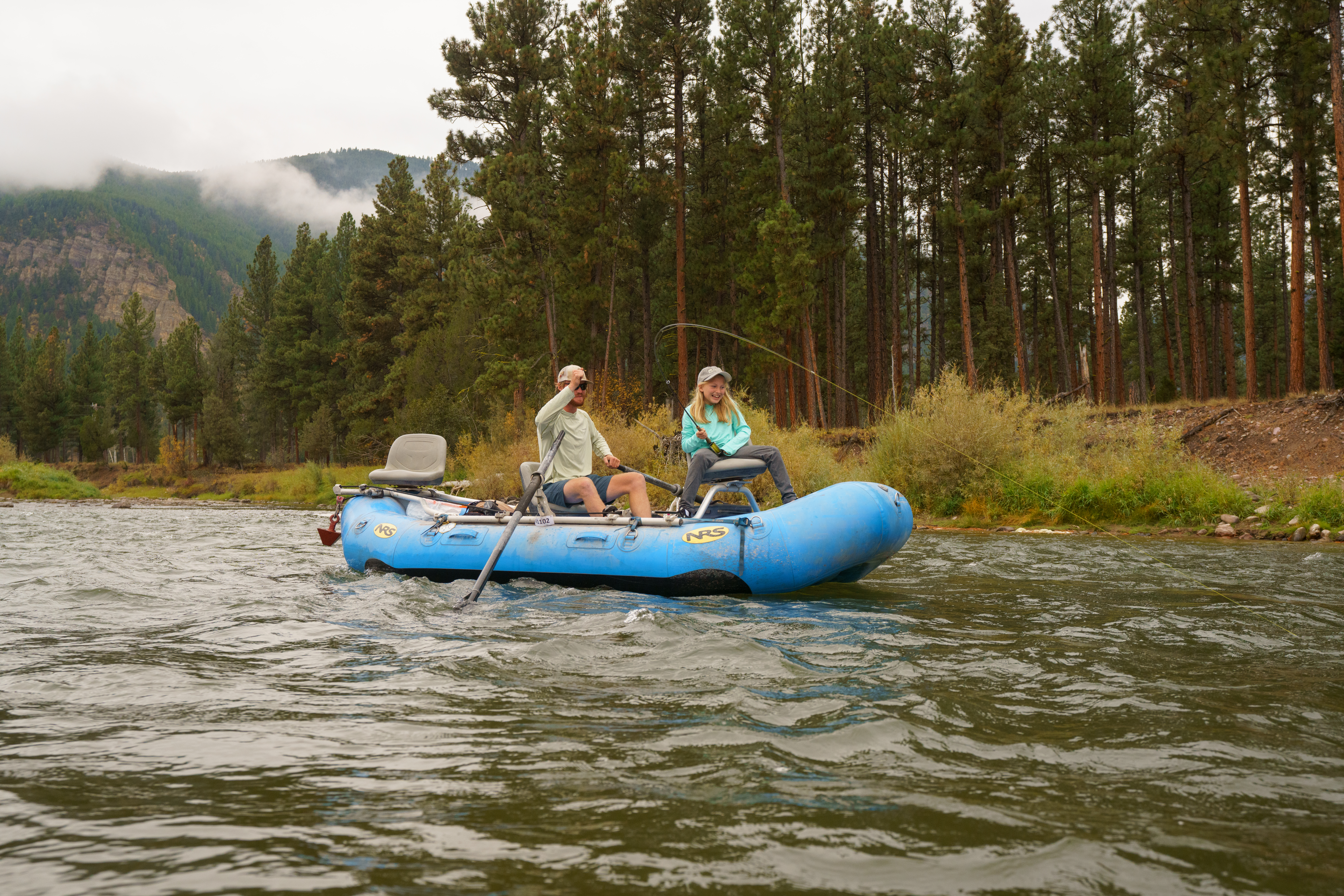 Man and young girl fishing in a blue boat with trees behind them in the distance.