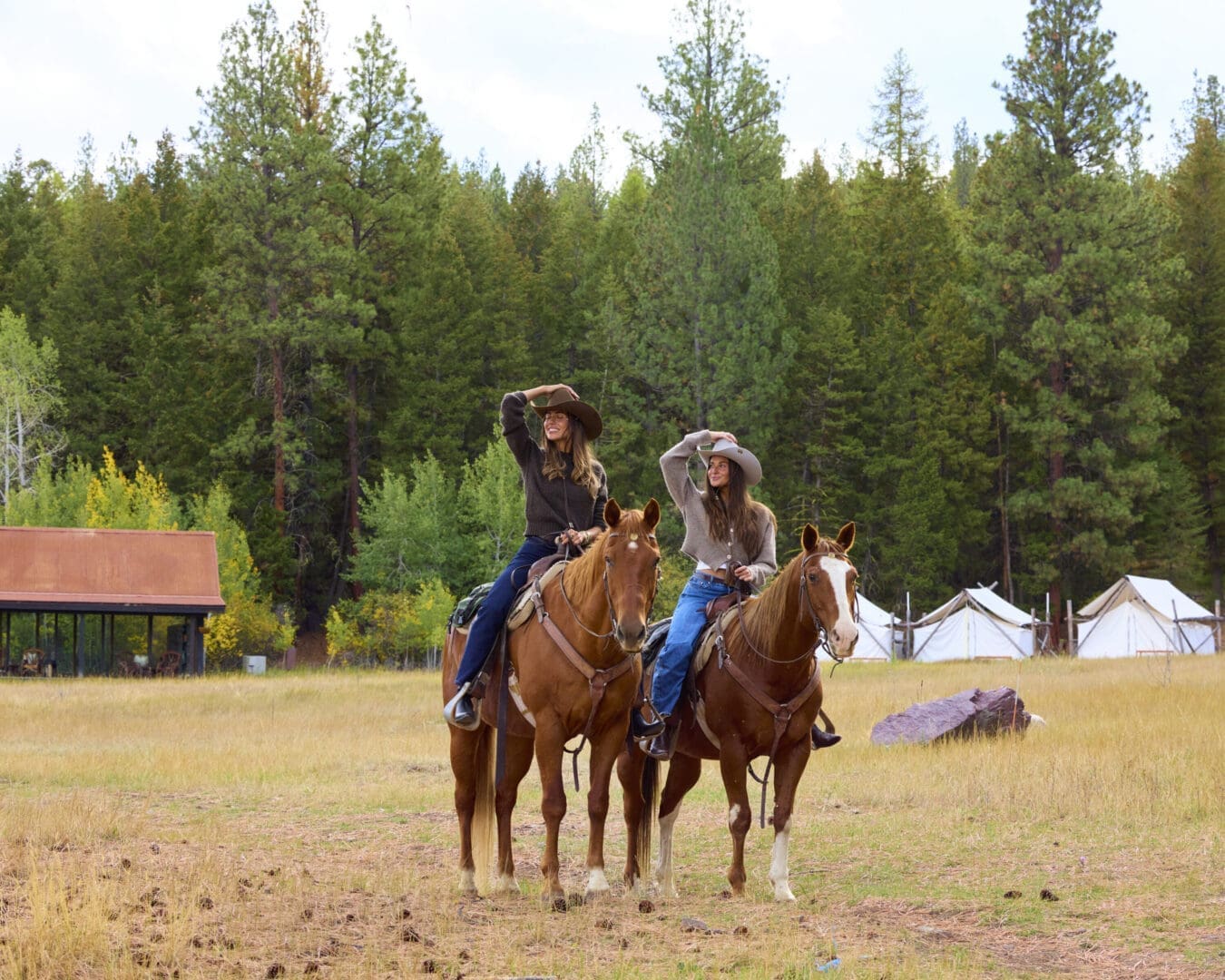 Two women on horseback with cowgirl hats surrounded by trees.