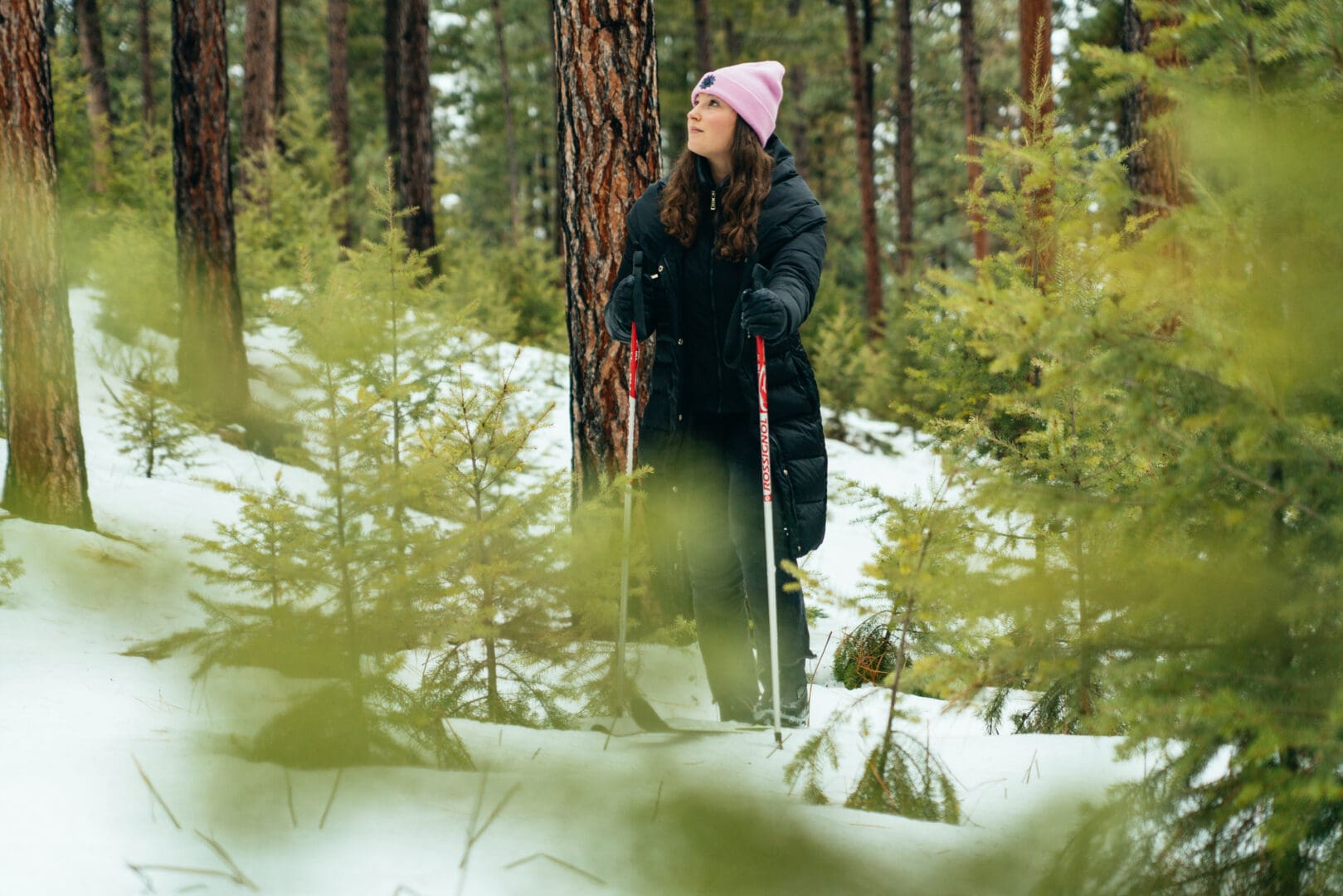 Woman wearing a black winter coat and pink hat outside in the snow with cross country skiis and poles.