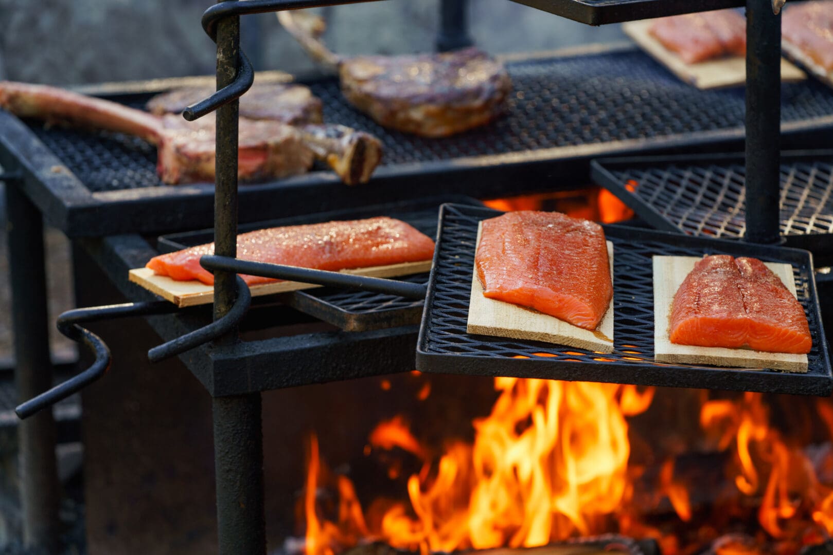 Salmon cooking over an open flame grill with steak tomahawks cooking in the background.