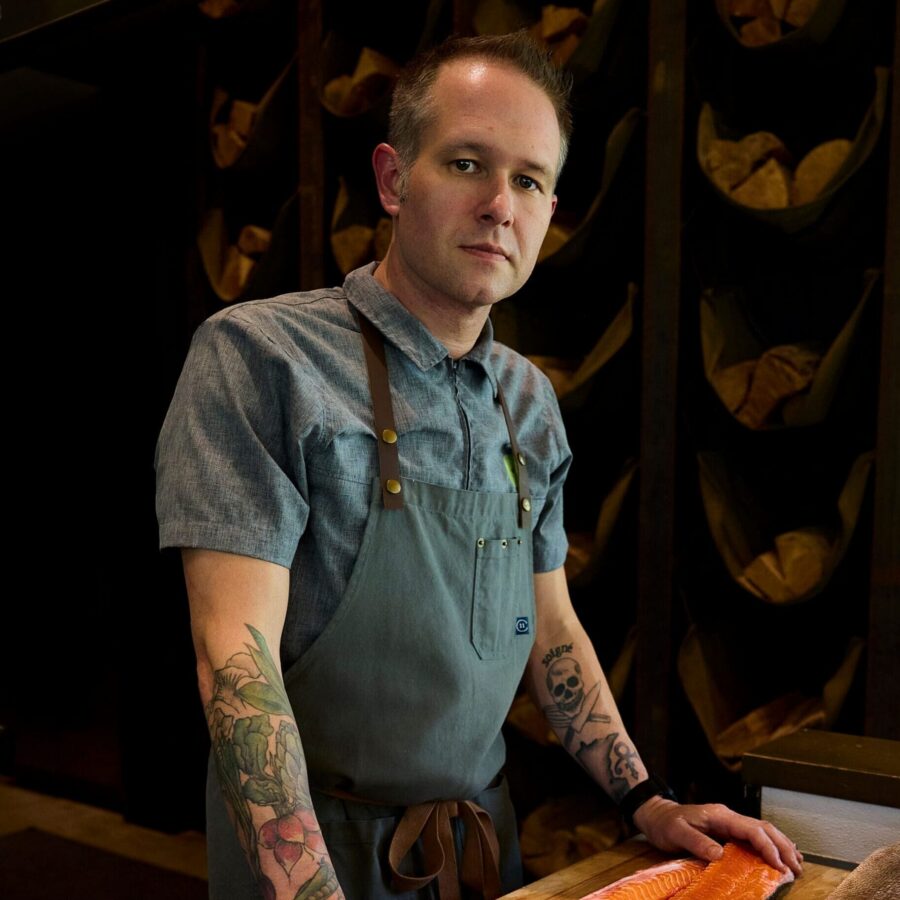 Chef standing next to cut up salmon on a cutting board in a dark elegant dining room.