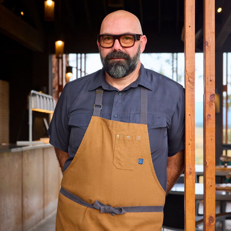 Chef posing in a tan apron in a wooden shed.