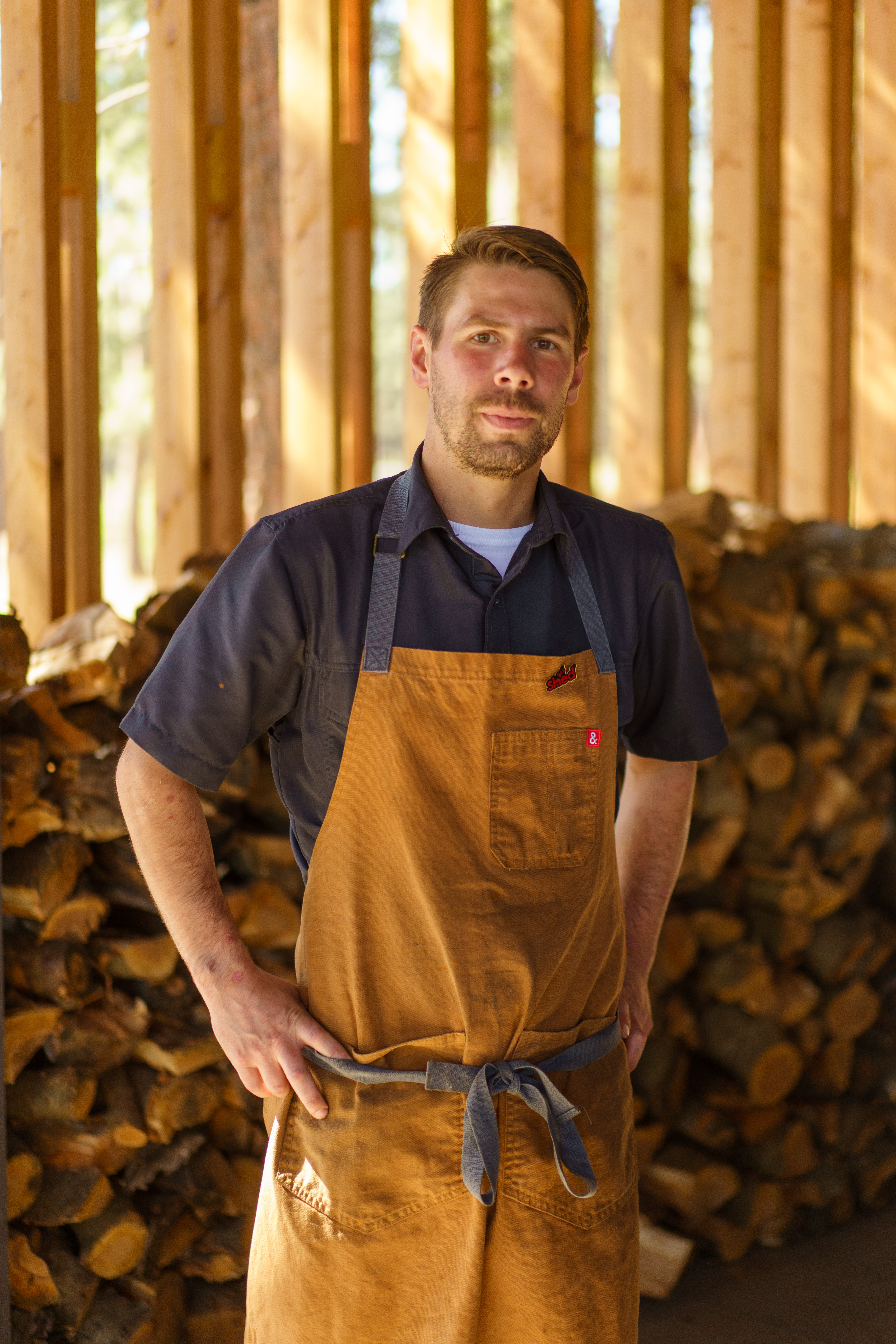 Chef posing in a tan apron in a shed with chopped wood behind him.