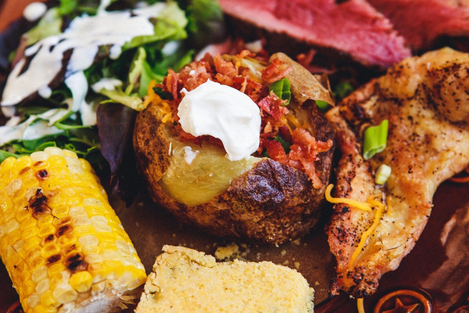 Closeup of grilled corn, salad, meat, and a stuffed baked potato topped with sour cream.