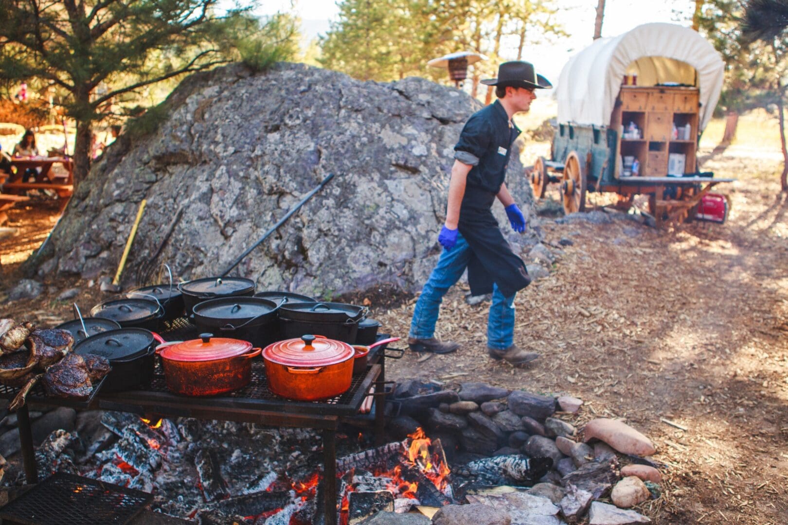 A chef wearing a cowboy hat walking away from attending to food cooking in red cast iron pots.
