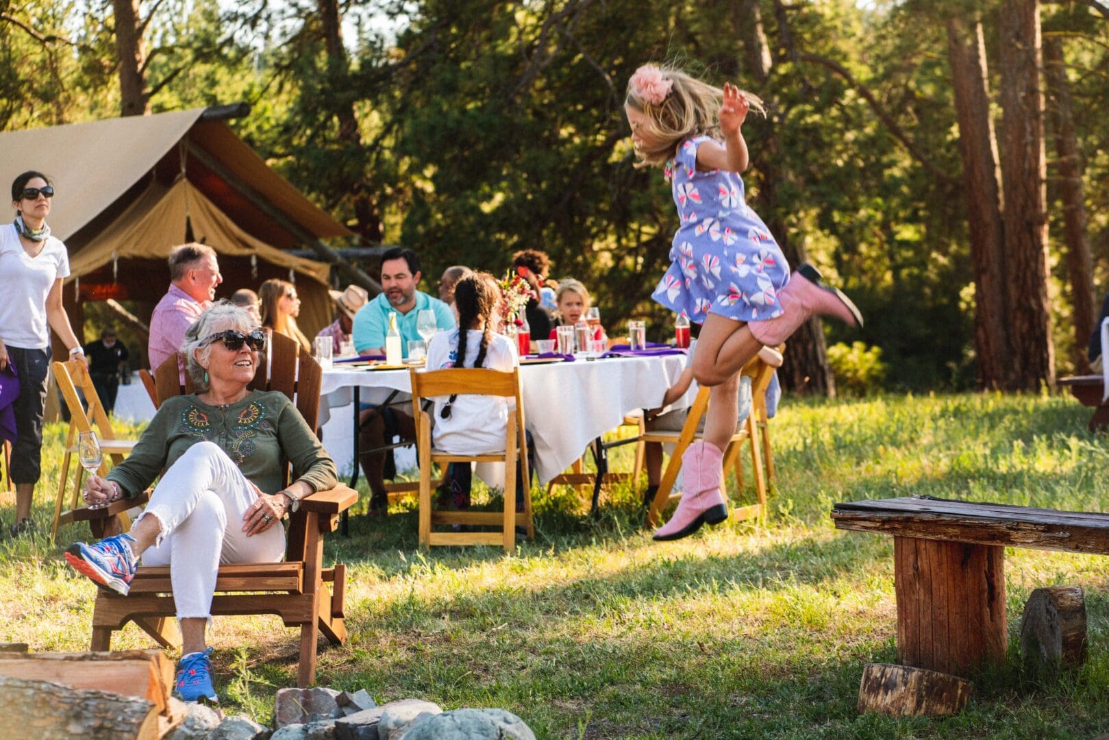Young girl jumping in the air while an elderly woman watches her, and a group of people are talking in the background at a picnic table.