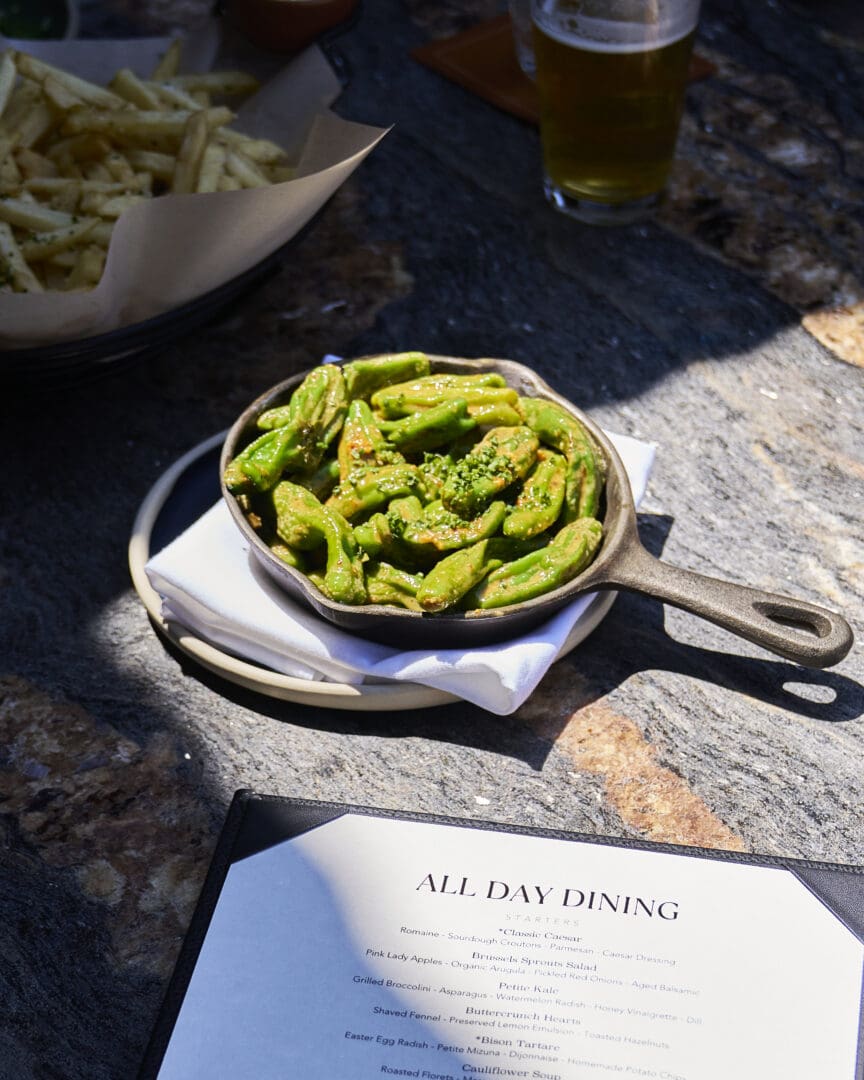 Green vegetables in a cast iron skillet sitting atop a wooden table outside with a fancy menu across from it.