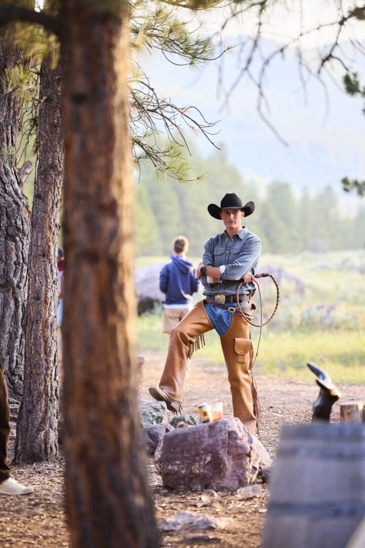 A cowboy in leather chaps standing and holding a lasso outside by trees.