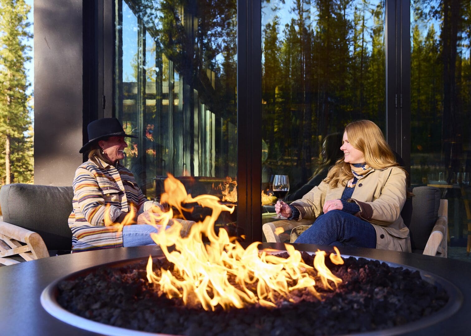 Two women sitting by a fire outside in front of a windowed building drinking wine.