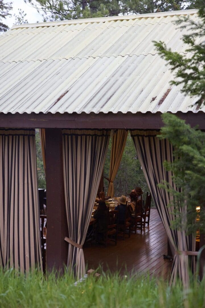 A pavilion with a tin roof and the curtains drawn back inside to show people sitting at a table indoors.