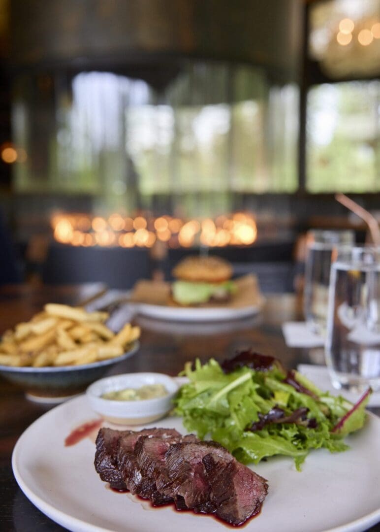 A dish of steak and salad on a table with a fire in a glass fireplace behind it.