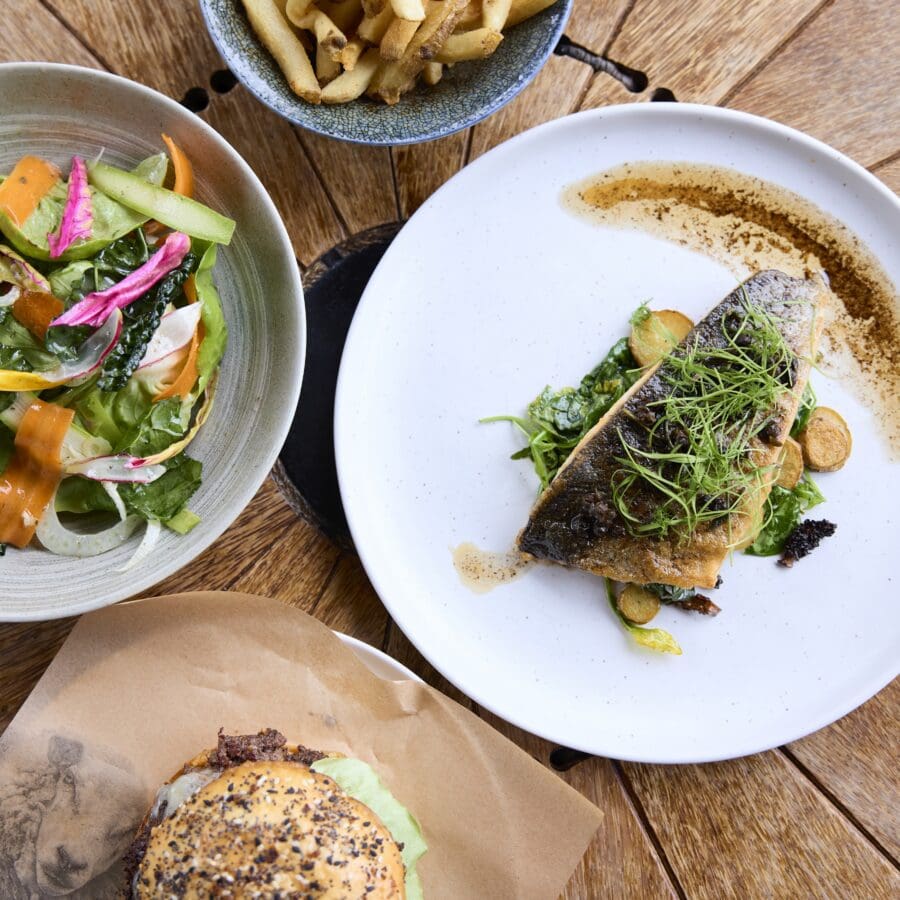 Four food dishes set out on a wooden table featuring salad, a burger, a fish dish, and fries.