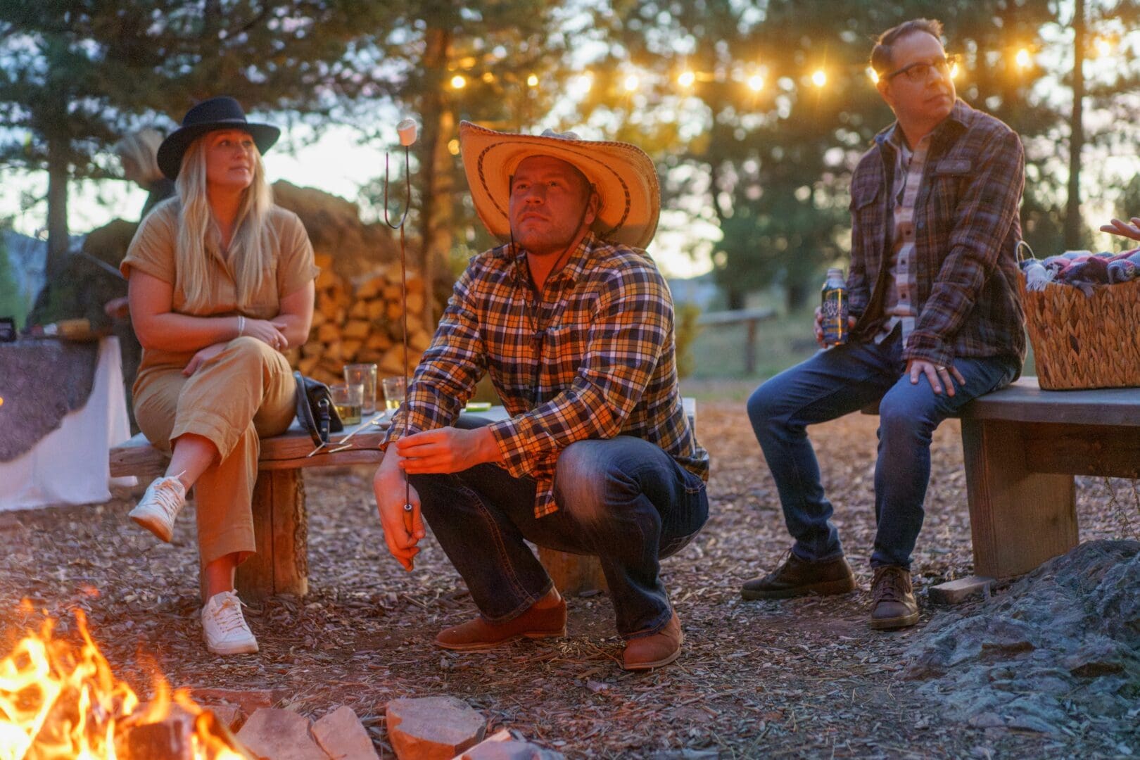 Three people sitting down by a fire with the middle man holding up a marshmallow roasting stick.