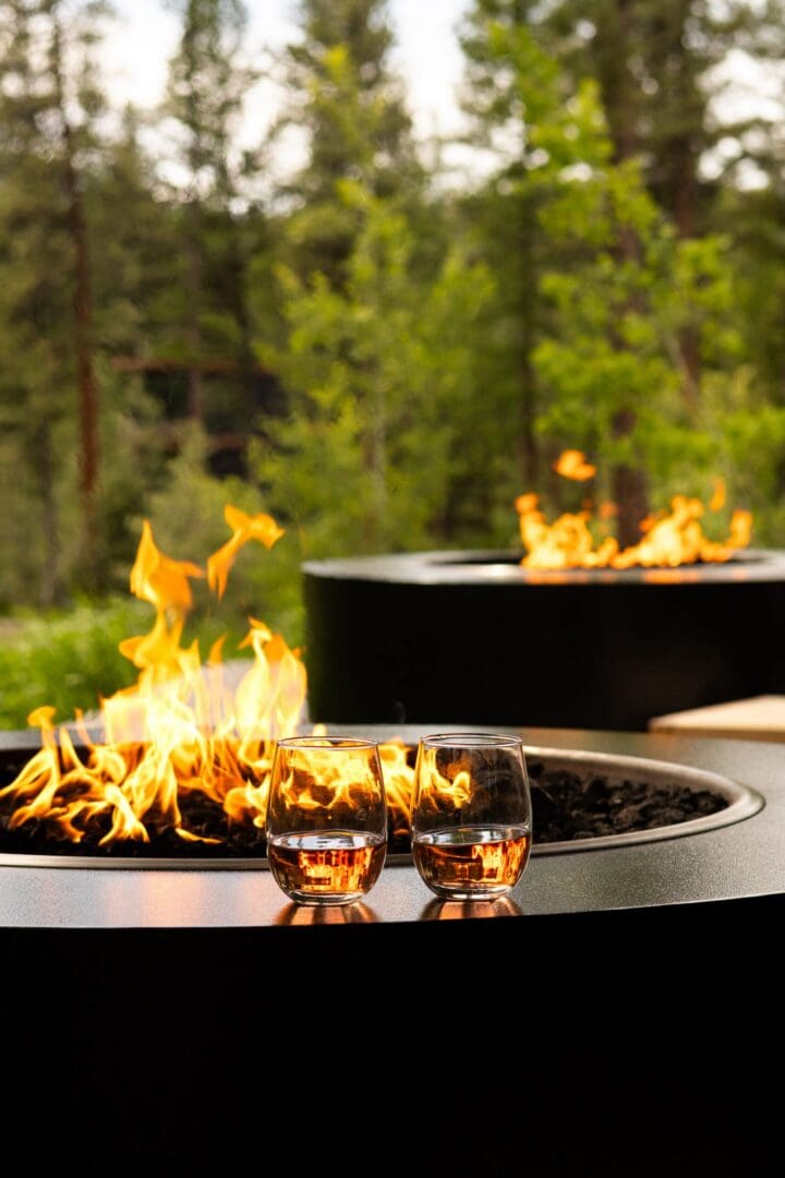 Two drinks sitting on the edge of a black fire pit with a live fire.