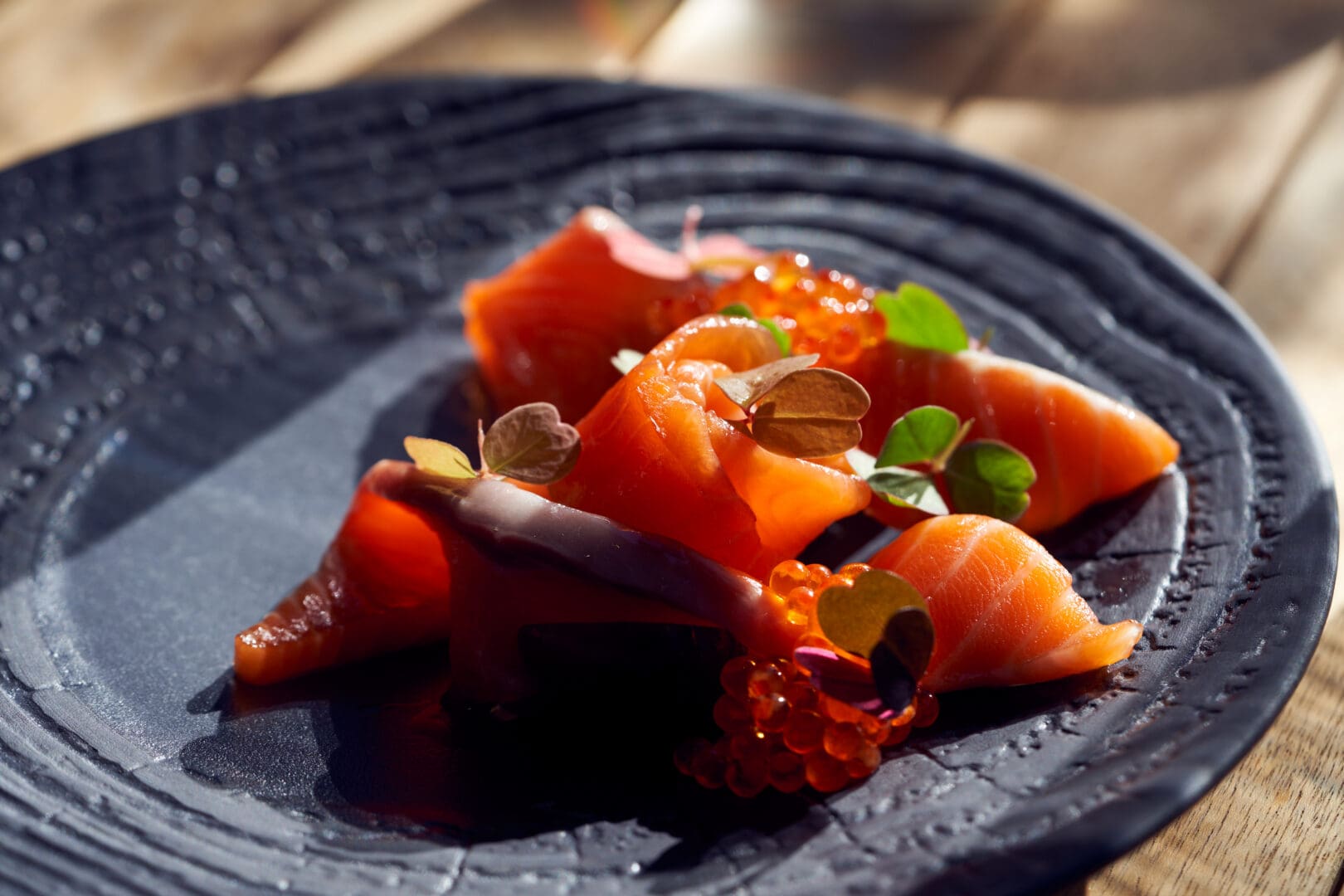 A salmon sliced fish dish with decorative greens on a dark plate.