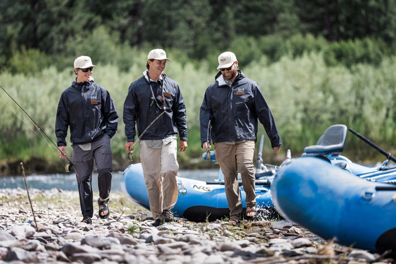 Three men walking away from blue rafts on a river carrying fishing poles.