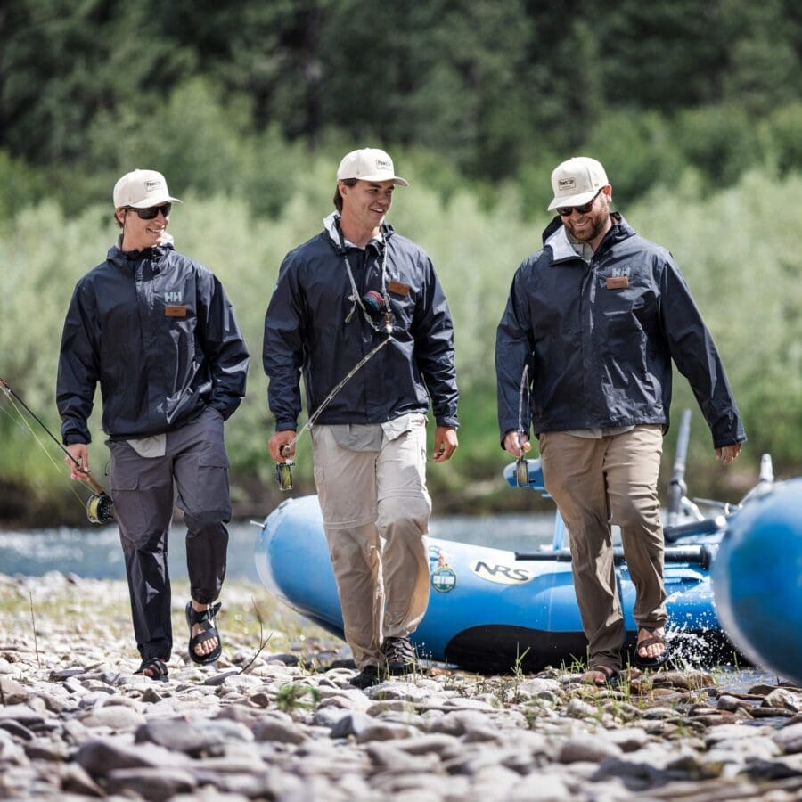 Three men walking away from blue rafts on a river carrying fishing poles.