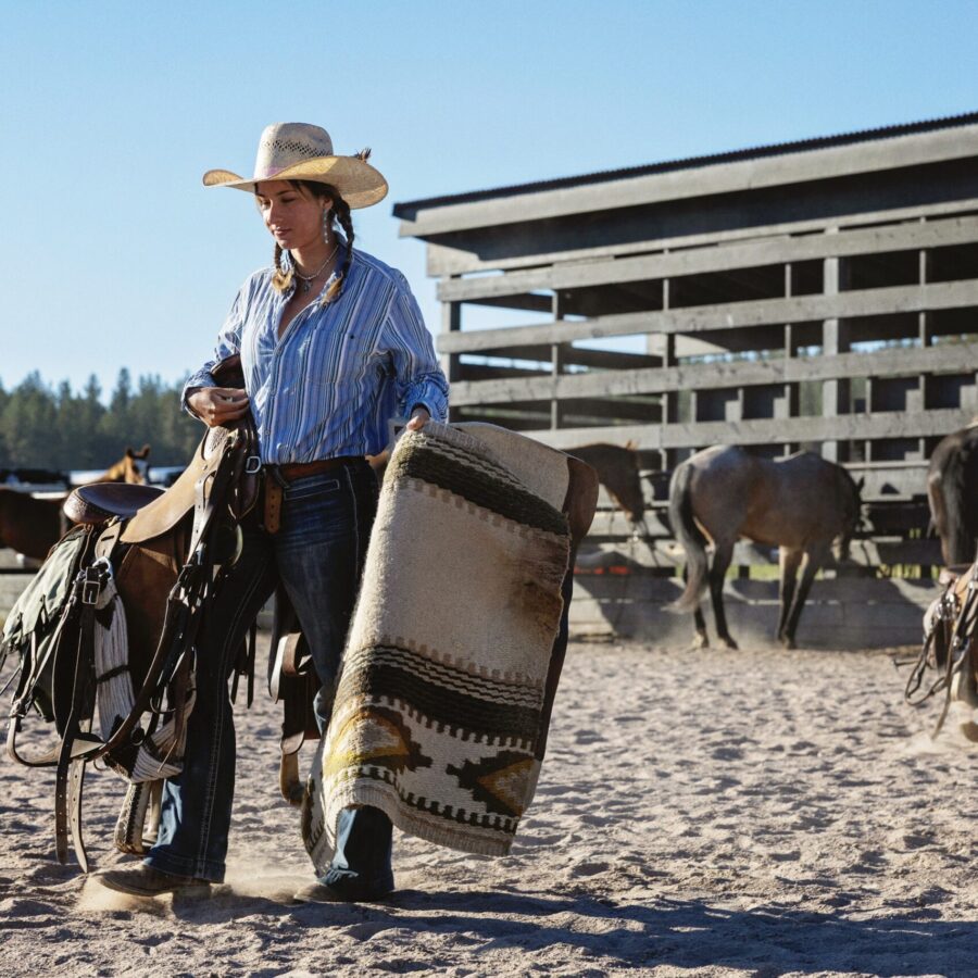 Wrangler carrying saddles in an outdoor arena.