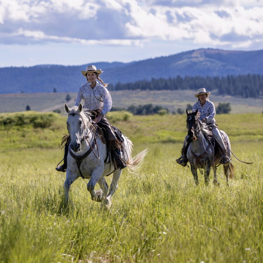 Two women riding horseback on a grassy field with mountains and cloudy skies behind them.