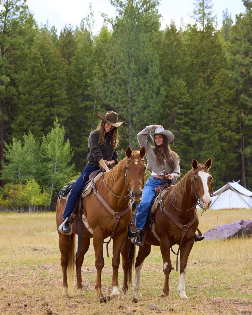 Two girls on brown horses with a green forest behind them.