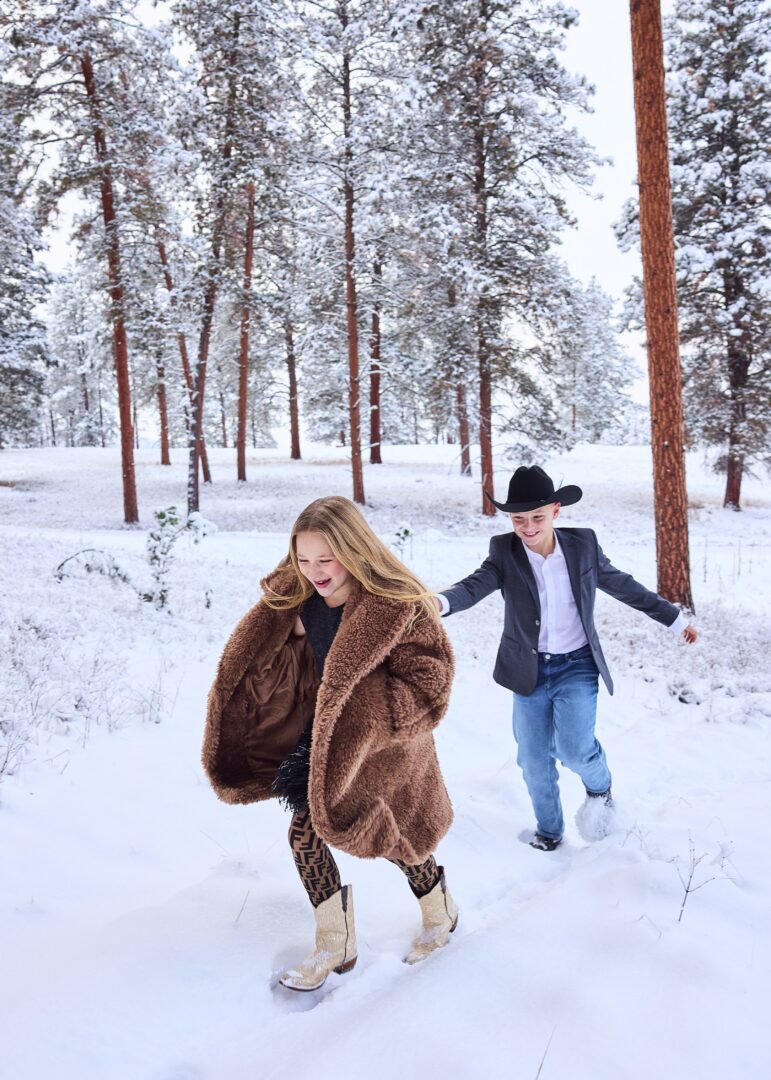 Two kids chasing each other through a snowy field with trees behind them.