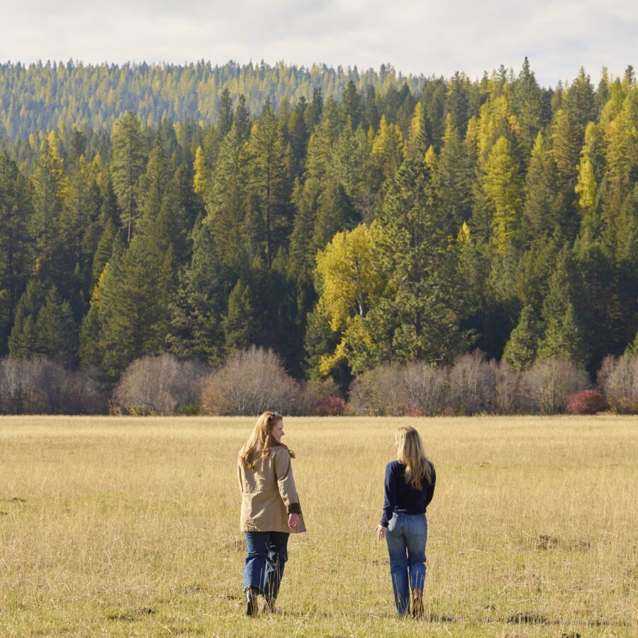 Two women walking on a field with colorful trees in front of them.
